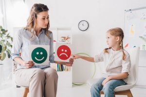 Child psychologist showing happy and sad emotion faces cards to child with autism spectrum disorder. Caregivers spend a lot of time in appointments.